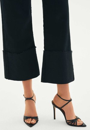 Sorte jeans med vide ben og frynset kant, kombineret med sorte højhælede sandaler med stropper, spids tå og twistdetalje.