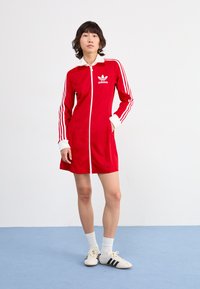 Rode sportieve jurk met een volledige rits, Adidas-logo en witte accenten. Heeft drie strepen op de mouwen en een contrasterende witte kraag.