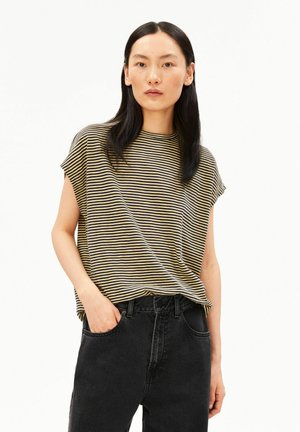 Femme aux longs cheveux foncés porte un haut beige et noir à manches courtes rayé, rentré dans un jean noir taille haute, une main dans la poche.
