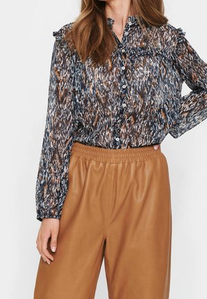 Nainen, jolla on läpikuultava, pitkähihainen kuviollinen paita, jossa on röyhelöitä, ja korkeavyötäröiset, vaaleanruskeat leveälahkeiset nahkahousut.