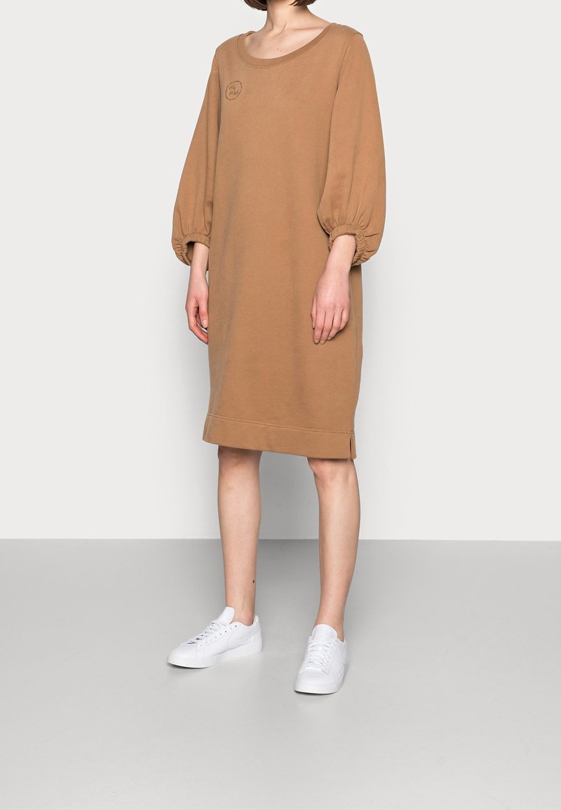 Hnědá oversized mikinová šaty s výstřihem do U, objemovými rukávy, žebrovanými manžetami a malým logem na hrudi. Nosené s bílými teniskami.