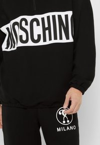 Černá mikina s bílým logem "MOSCHINO" přes prsa a odpovídající černé kalhoty s bílou grafikou "MILANO".