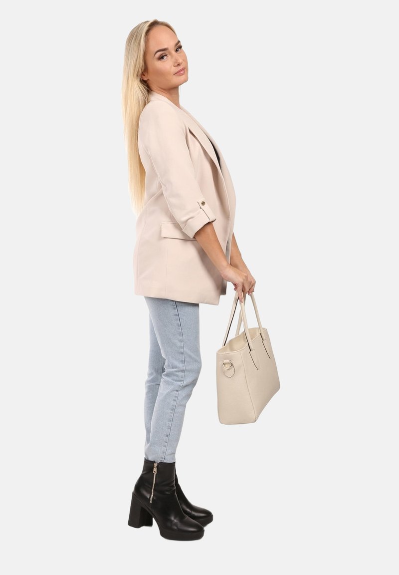 Lichtbeige blazer met opgerolde mouwen, gecombineerd met lichtblauwe jeans en zwarte enkellaarsjes met hak, en houdt een lichtgrijze handtas vast.