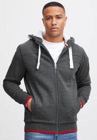 Grijze rits-hoodie met een witte fleece-voering in de capuchon, rode accenten aan de zoom en witte koordjes. Glad textuur, geribbelde manchetten en zakken.