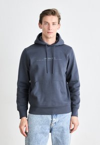 Donkergrijze fleece hoodie met een buidelzak, ribgebreide manchetten en een witte tekstlogo over de borst. Gecombineerd met lichtblauwe jeans.