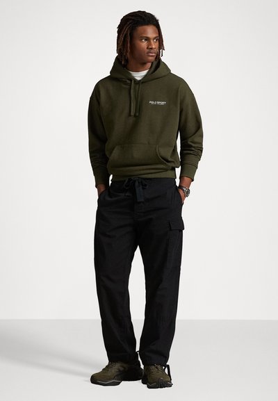 Hanorac verde închis cu buzunar frontal și logo, purtat cu pantaloni cargo negri care au buzunare laterale. Încălțăminte sport verde închis.