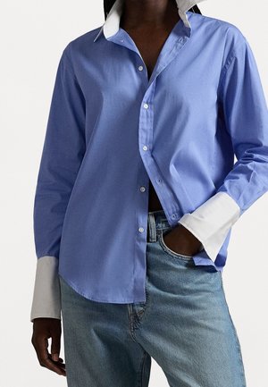 Persoon draagt een gedeeltelijk opengeknipt blauw shirt met witte kraag en manchetten, gecombineerd met lichtblauwe jeans, hand in de zak.