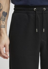 Zwarte trainingsbroek gemaakt van zachte, gestructureerde stof. Beschikt over een elastische tailleband en een trekkoord met metalen uiteinden. Zijdelingse zakken zijn zichtbaar.
