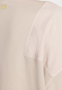 Beiges T-Shirt mit einem glatten Baumwollkörper und einem kontrastierenden, strukturierten Stoffpaneel an der Schulter. Goldene Schrift auf der Rückseite.