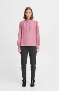 Langärmlige pinkfarbene Bluse mit Rüscheneinsatz an der Vorderseite und hohem Kragen, kombiniert mit dunkelgrauen, schmal geschnittenen Hosen und schwarzen Ankle Boots.