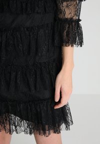 Svart blondekjole med volanger, som har transparente ermer og et teksturert, mønstret stoff. Nærbilde viser intrikate detaljer og en hånd.