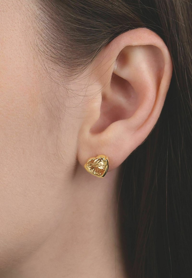 Nahaufnahme eines Frauenohres mit einem goldenen, herzförmigen Ohrstecker mit einem facettierten gelben Edelstein.