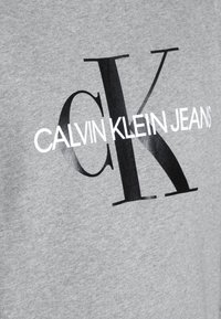 Šedá látka s velkými černými písmeny "CK" a bílým textem "CALVIN KLEIN JEANS" vytištěným diagonálně přes střed.