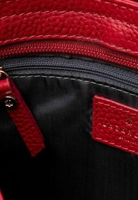 Interior de un bolso de mano de cuero rojo con un acabado texturizado, que cuenta con un forro oscuro, un bolsillo con cremallera y una etiqueta de la marca grabada en rojo.