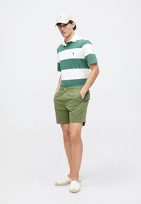 Polo a righe verde e bianche, pantaloni corti khaki e espadrillas bianche. Gli accessori includono un cappello chiaro. Outfit casual su un modello in piedi.