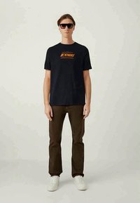 Muž oblečený v černém tričku K-Way, hnědých kalhotách, bílých teniskách a černých slunečních brýlích, stojící na pozadí v jednobarevném světle.