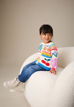Joven chica con un suéter de colores con motivos y vaqueros azules sentada en un sillón blanco texturizado, mirando de lado con una ligera sonrisa.