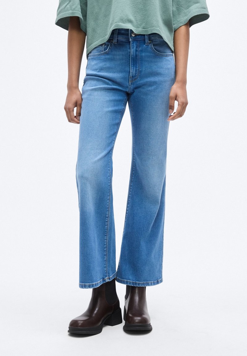 Personne portant un jean large bleu clair, un haut vert ample et des bottines à talons brun foncé, debout devant un fond uni.