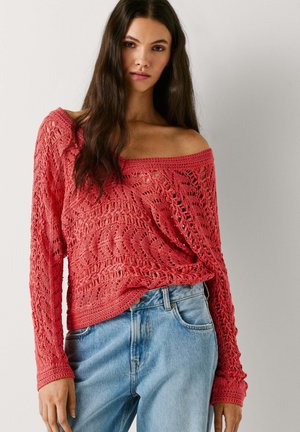 Mlada ženska z dolgimi temnimi lasmi, oblečena v koralno rdeč pulover z odkritimi rameni iz kvačkanega materiala in svetlo modre kavbojke, stoji ob navadni steni.