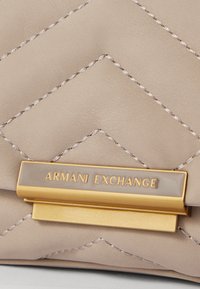 Cremefarvet, kviltet, kunstlæder håndtaske med en guldfarvet lås, der har "Armani Exchange" i hævet skrift. Teksturering med syninger.