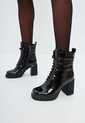 Bottines en cuir verni noir avec un haut talon carré, un devant lacé, une fermeture éclair sur le côté et une finition texturée. Elles présentent un bout arrondi et des lacets ajustables.
