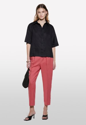 Schwarzes kurzärmeliges Button-up-Hemd, lockere Passform, kombiniert mit rosa maßgeschneiderten Hosen. Model hält eine schwarze geflochtene Handtasche und trägt schwarze offene Absatzschuhe.