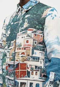 Chaqueta con cremallera que presenta un estampado detallado y colorido de edificios costeros y vegetación sobre tela.