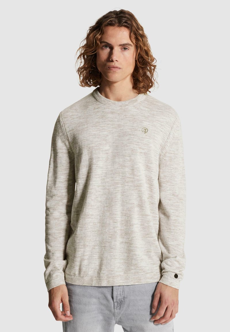 Jeune homme aux cheveux bouclés mi-longs portant un pull gris clair à manches longues et un pantalon gris clair sur un fond uni.