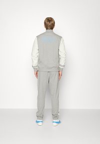 Grå Nike-jacka med vita ärmar, ljusblå logotyp på ryggen, tillsammans med matchande grå byxor och sneakers med blå accenter.