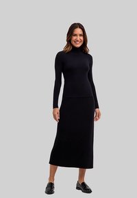 Zwarte ribgebreide jurk met lange mouwen, hoge hals en een rok tot op de knie. Draagt zwarte loafers. Gladde textuur en aangesloten ontwerp.