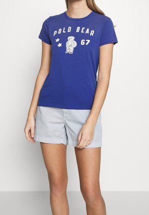 Femme portant un t-shirt bleu avec le texte « Polo Bear 67 » et un graphique d'ours, associé à un short gris clair, debout devant un fond blanc.