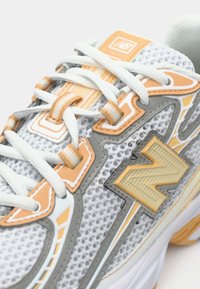 Pogled od blizu na bele in oranžne superge New Balance z mrežasto tkanino, belimi vezalkami in velikim logotipom "N" na strani.