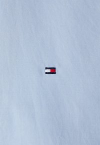 Tessuto blu chiaro con un piccolo logo ricamato che presenta elementi rossi, bianchi e blu navy, situato al centro a sinistra del materiale.