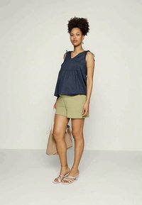 Top sem mangas azul-marinho com decote em V e cintura franzida, combinado com calções verde-claro e sandálias rasas. Bolsa de toalha bege sobre um ombro.