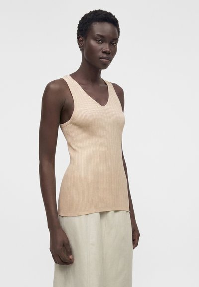 Γυναίκα με αμάνικη μπεζ ριμπ πλεκτή μπλούζα και ανοιχτόχρωμη φούστα, στέκεται μπροστά σε απλό φωτεινό φόντο.