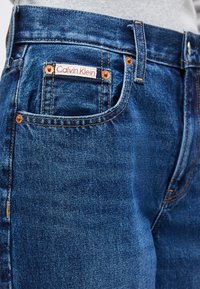 Κοντινό πλάνο τσέπης γαλάζιου τζιν με χάλκινα καρφιά και μια μικρή λευκή ετικέτα με την επιγραφή "Calvin Klein" ραμμένη πάνω από την τσέπη.