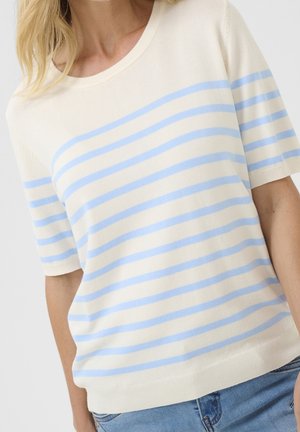 Vrouw draagt een kort mouw crème trui met lichtblauwe horizontale strepen en een blauwe spijkerbroek tegen een effen achtergrond.