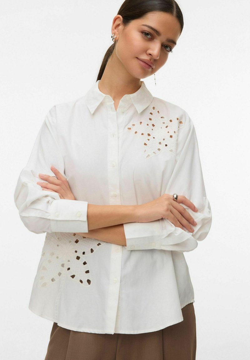 Chemise blanche boutonnée avec motif floral découpé sur le côté gauche, dotée d'un col classique et de manches longues. Tissu en coton lisse.