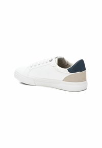 Zapatilla blanca con un upper sintético liso, punta redondeada y forro textil. Presenta un acento beige y azul oscuro en el talón.