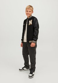Svart varsityjacka med lädersleeves, randiga manschetter och ett guld broderat bokstav "N." Bärs över en ljus skjorta med mörka jeans och sneakers.