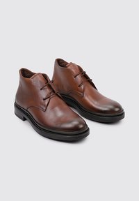 Botas de cuero marrón hasta el tobillo con una textura suave, punta redondeada y diseño minimalista. Presenta cuatro ojales y una suela de goma negra.