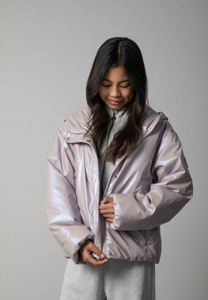 Młoda kobieta o długich ciemnych włosach zapinająca błyszczącą, jasnofioletową kurtkę na szarym swetrze i spodniach na jednolitym szarym tle.