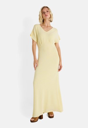 Robe maxi rayée jaune et blanche avec un décolleté en V, des manches courtes à volants et un léger drapé au niveau de la poitrine. Portée avec des chaussures à plateforme marron.