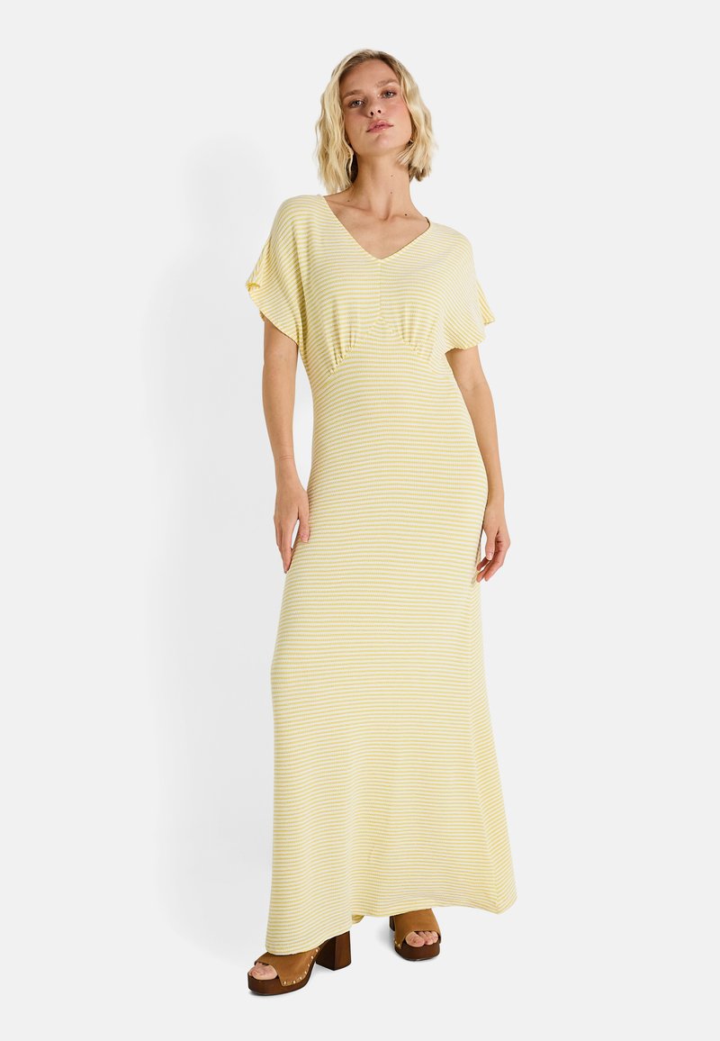 Robe maxi rayée jaune et blanche avec un décolleté en V, des manches courtes à volants et un léger drapé au niveau de la poitrine. Portée avec des chaussures à plateforme marron.