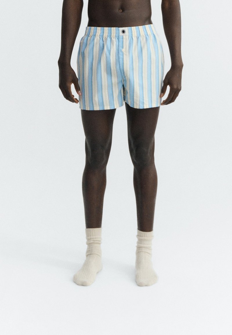 Homme debout portant un boxer à rayures verticales bleues et blanches avec des chaussettes blanches, sur un fond uni.