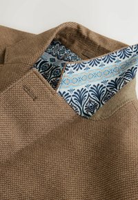 Col de blazer marron texturé relevé pour révéler la doublure en soie bleue et blanche à motifs floraux et géométriques.