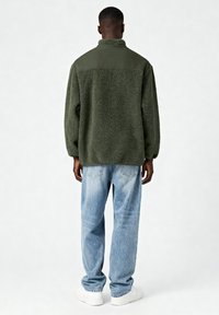 Veste en polaire verte avec un col haut et un corps texturé, associée à un jean bleu clair et des baskets blanches. Coupe décontractée, design relax.