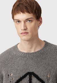 Giovane con capelli corti castani che indossa un maglione grigio a maglia con un simbolo di pace nero e piccoli buchi consumati.