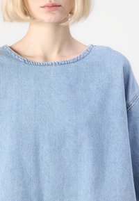 Top in denim azzurro chiaro con scollatura ampia, silhouette informale e maniche leggermente accorciate. Presenta cuciture visibili lungo i bordi.