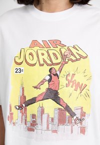 Hvid t-shirt med et grafisk tryk af en springende spiller, "AIR JORDAN" tekst i gul, med byens skyline i baggrunden og tegneserie-lignende effekter.